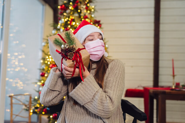 惊喜新年前夜拿着圣诞礼物的女孩看着相机的女孩冠状病毒期间的圣诞节 概念闪亮松树检疫