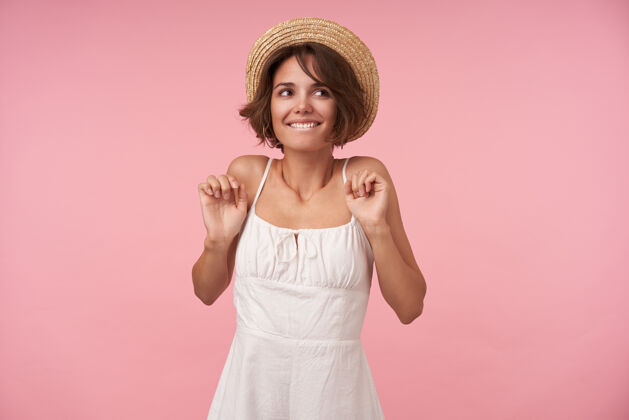 深色一个快乐的黑发女人 她剪着短发 一边积极地看着一边 一边咬着内衣 一边穿着白裙子 戴着草帽 一边举手摆姿势夏天帽子可爱