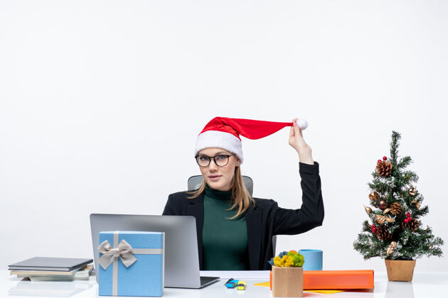 圣诞节一个积极的女商人坐在一张桌子旁 手里拿着圣诞树和礼物 手里拿着一顶圣诞老人的帽子 在白色的背景下查看邮件商务女人玩圣诞老人