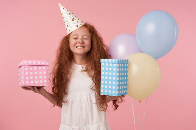 空气快乐的红发小女孩 长卷发 白色连衣裙 戴着生日帽庆祝节日 手里拿着礼物 脸上洋溢着喜悦的笑容 在粉色的工作室背景下与世隔绝孩子高兴室内