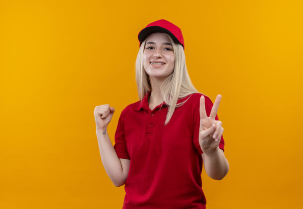 送货微笑的小女孩 穿着红色t恤 戴着帽子 戴着牙套 在孤立的橙色背景下 表现出平和和和的姿态和平年轻帽子