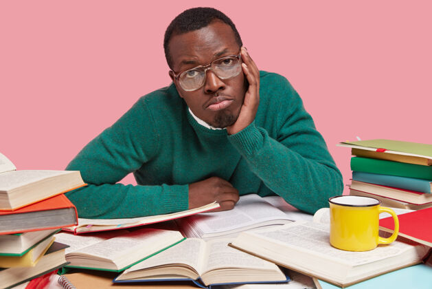 美心烦意乱的黑皮肤男人有着无聊的表情 手放在脸颊上 戴着眼镜 穿着绿色毛衣 身边围着许多书大学嘴书桌