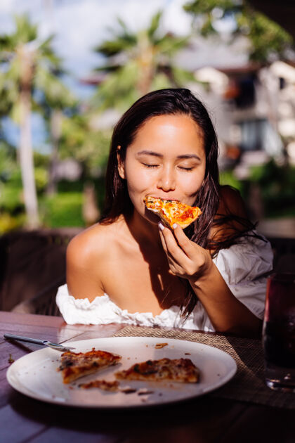 笑快乐的亚洲美女在阳光明媚的日子里饿着肚子吃披萨在户外餐厅日落的阳光下女人在享受美食在午餐的乐趣女性快乐户外