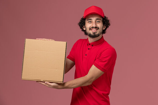 工作正面图：身穿红色衬衫和披风的男性快递员手持浅粉色墙上的送货食品箱微笑男性快递成人