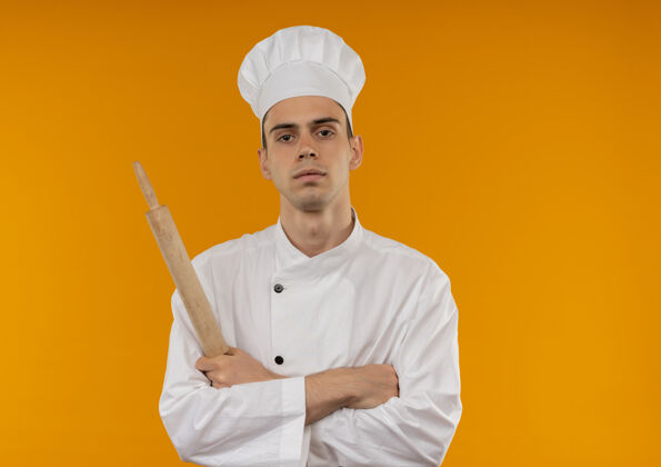 看年轻男酷穿厨师制服交叉手拿擀面杖在孤立的黄色墙壁与复制空间年轻酷交叉