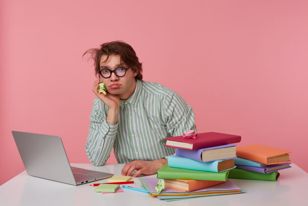 笔记本疲惫的年轻黑发男性摆姿势 穿着条纹衬衫 戴着眼镜 坐在工作桌旁 拿着笔记本电脑和书 头靠在手上 神情憔悴准备男人深色