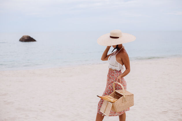 热带沙滩上的浪漫女人穿着裙子 针织上衣 戴着草帽 抱着面包篮子 生态生活浪漫面包度假