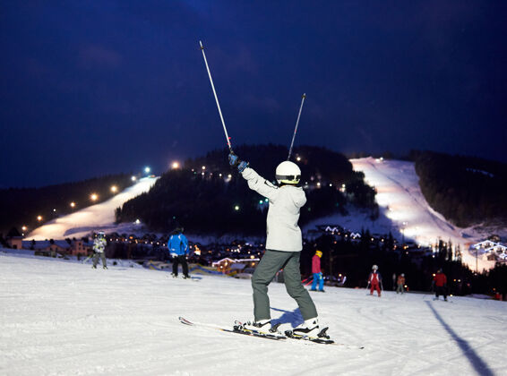 娱乐手举滑雪竿的无法辨认的女滑雪者后视图深蓝的天空下 树木繁茂的山上有滑雪道自然喀尔巴阡山夜晚