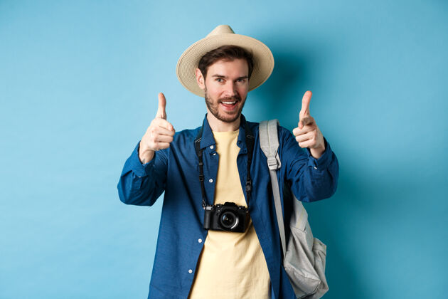 帽子帅气的游客带着相机和背包去度暑假 站在蓝色的背景上 手指着相机 邀请着你喜欢站胡子