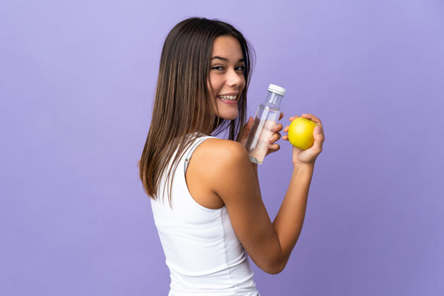 美味一个女人用一个苹果和一瓶水隔离在紫色的土地上液体女人矿物质