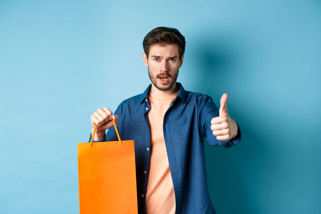 酷帅哥竖起大拇指 拿着橙色的购物袋 站在蓝色的背景上情感年轻肖像