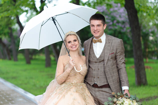 下雨快乐的新郎新娘打着白色雨伞 在夏天的公园里露天婚礼新娘和新郎拥抱妻子