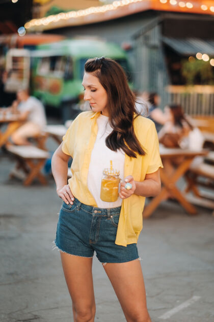 时尚一个穿着黄色衬衫和短裤 喝着一杯柠檬汁的女孩站在街上新鲜女人街头