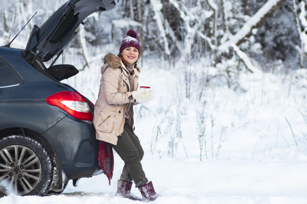 舒适一个舒适的女人微笑着 一个穿着温暖冬衣的女人喝着热饮 茶或咖啡 坐在汽车后备箱里微笑着度假 开车旅行 下雪寒冷复制空间微笑周末冬天