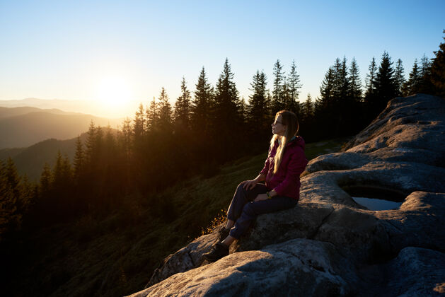 日落日落时分坐在山上巨石上的女徒步旅行者黎明享受早晨