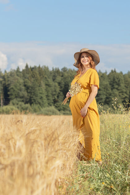 农场在一个阳光明媚的夏日 一位年轻漂亮的孕妇穿着黄色的裙子 戴着黄色的帽子 走过一片麦田风景收获农村