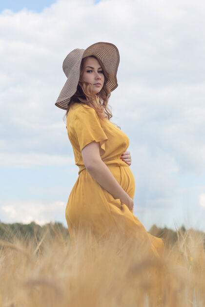 帽子在一个阳光明媚的夏日 一位年轻漂亮的孕妇穿着黄色的裙子 戴着黄色的帽子 走过一片麦田阳光收获天空
