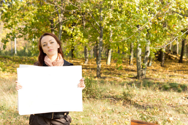 自然美丽的年轻女子站在树木繁茂的乡间 手里拿着一张白卡年轻展示女人