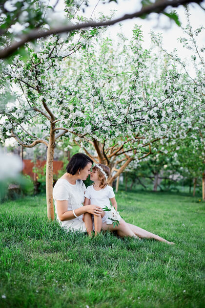 欢笑可爱的女儿和妈妈坐在花开的春天花园里 抱着快乐的女人和孩子 穿着白色的衣服在户外 春天的季节即将到来母亲节假期概念复活节幼儿玩耍
