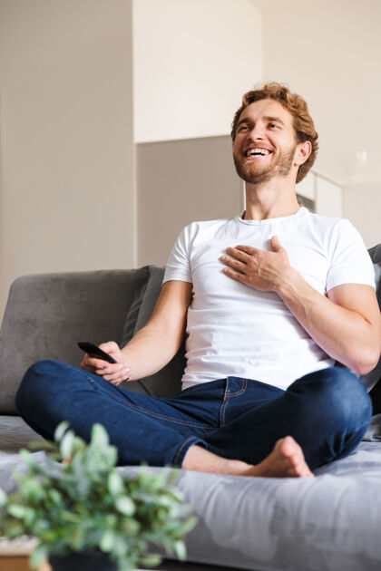 舒适一个英俊快乐的年轻人在家里的沙发上拿着遥控器看电视放松休闲控制