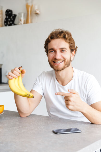 学习照片中 一个快乐的留着胡子的年轻人在家里的餐桌上指着香蕉和水果舒适教育白种人