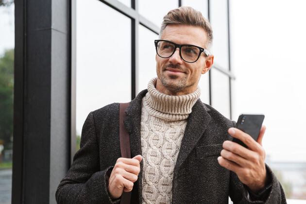 聊天30多岁的时尚男人戴着眼镜 穿过城市街道 使用智能手机的画像帅气休闲城市
