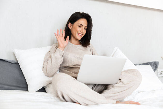消遣图为30多岁的快乐女人在舒适的公寓里用笔记本电脑 穿着白色亚麻布坐在床上公寓沙发舒适