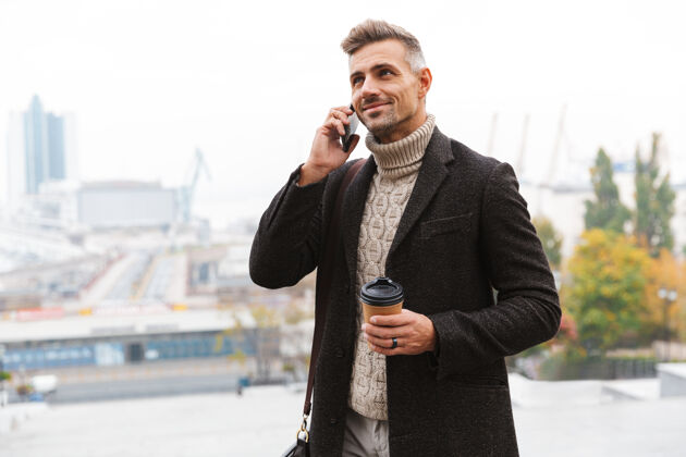 成人一张30多岁的时尚男士穿着夹克 一边在城市街道上散步 一边用手机聊天 一边拿着外卖咖啡的照片城镇户外手持