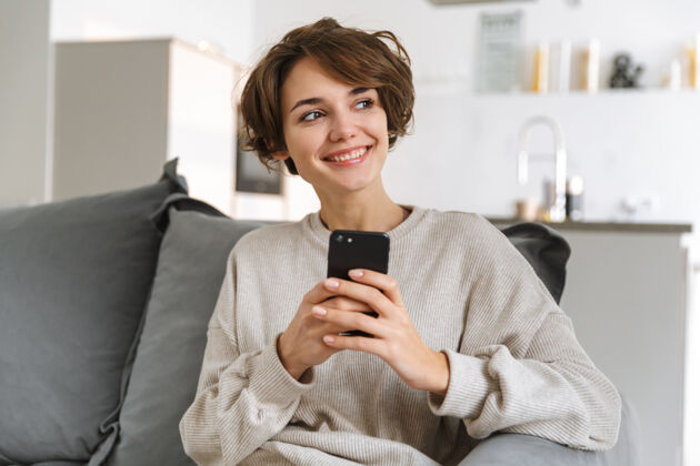 手机快乐的年轻女子坐在家里的沙发上 用手机沙发女士应用程序