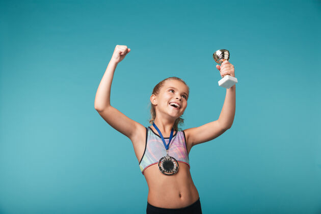 成功快乐的小体育女孩庆祝战胜蓝墙 戴着金牌 展示着奖杯小比赛最佳