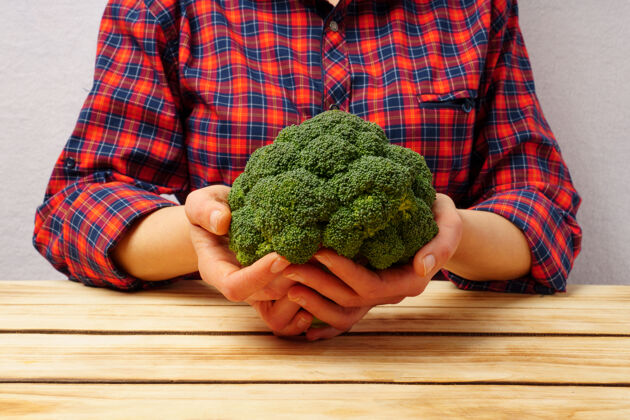 素食绿色的新鲜西兰花在女性手中 身着红色格子衬衫的木桌背景上沙拉生的食物