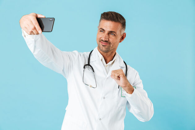 服务穿着白大褂 手持听诊器的成熟医生在手机上自拍的照片 孤零零地站在蓝色的墙上制服救护车手机