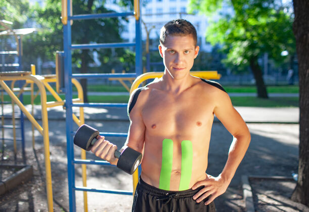 比赛帅气的年轻白种人职业健美运动员的肖像 身上带着黑色和绿色的松紧带 在运动场上举着哑铃哑铃锻炼锻炼