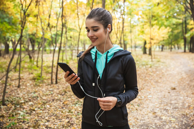 电话微笑的健身女 戴着耳机听音乐 手持手机站在公园里成人跑步女运动员