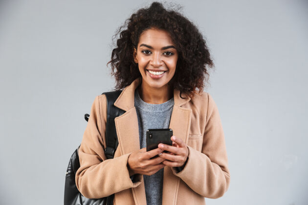 通话微笑的非洲年轻女子穿着外套 一边用手机 一边孤立地站着设备衣服手机