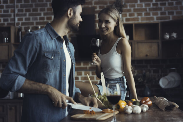 欢呼美丽的年轻夫妇在家里的厨房边喝酒边微笑准备木头食谱