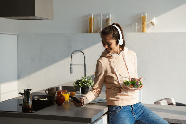 早餐快乐的年轻女孩在家的厨房里用耳机听音乐 吃着碗里的沙拉成人休闲放松