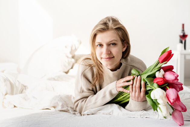 人幸福的年轻女子穿着睡衣躺在床上捧着郁金香花束微笑新鲜庆祝