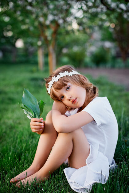 复活节可爱的5-6岁抱着花的小女孩坐在百花齐放的春天花园里 穿着白色的连衣裙 戴着花环在户外 春天来了美丽休闲小