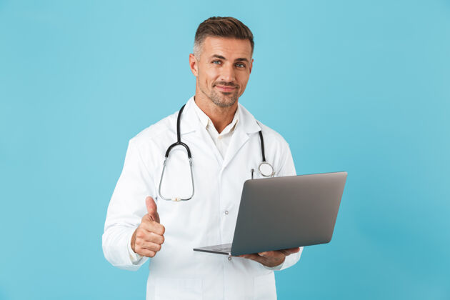 从业者照片中的成年男子身穿白色医用外套 手持手提电脑的听诊器 孤立地站在蓝色的墙上东西积极男人