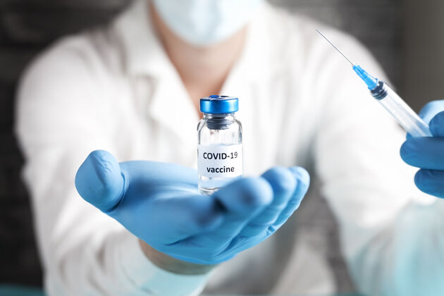药房研究人员手里拿着Covid-19疫苗 男医生穿着白大褂 戴着医用口罩 戴着蓝色手套 手里拿着装有治疗冠状病毒疫苗的注射器和瓶子冠状病毒治疗的概念玻璃测试患者