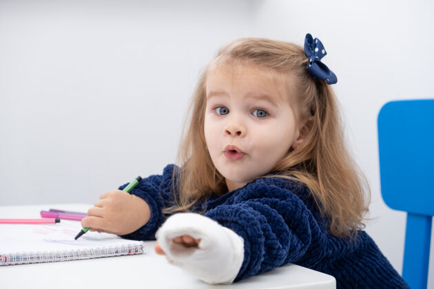 治疗坐在桌边用马克笔试着画画的小女孩手拿石膏矫形绷带创伤
