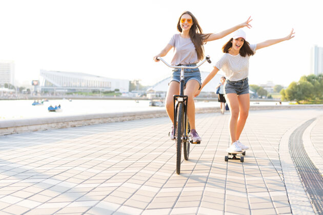 阳光两个年轻的朋友在一个炎热的夏日放松 一个年轻的女人骑着自行车拖着她的朋友在一条城市街道上滑滑板城市女朋友城市