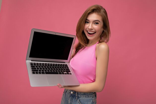 游戏幸福的微笑美丽的年轻金发女人拿着电脑笔记本穿着粉色的短上衣看着相机隔离在粉红色背景.mock起来 复制空间乐趣社交技术