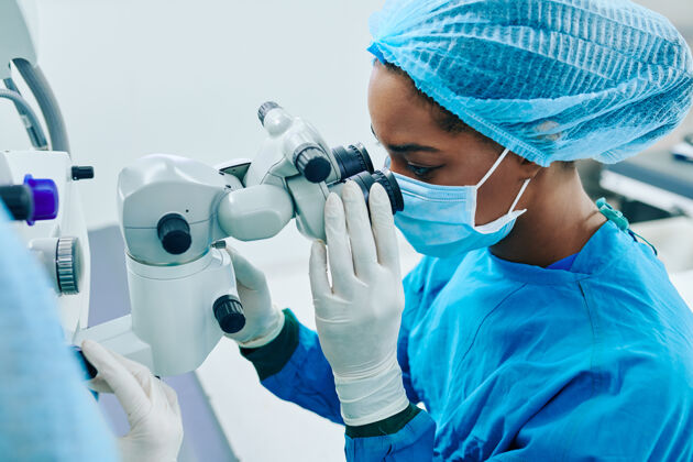 体检年轻的黑人女外科医生戴着医用面罩和橡胶手套透过电子显微镜看工作外科医生医疗职业