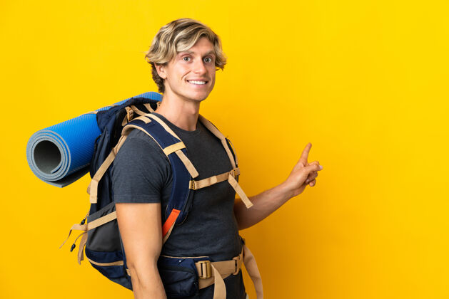 金发年轻的登山者在孤立的黄色背景上指着后面背包美国主持人