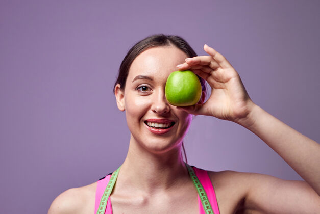 锻炼年轻漂亮的运动女孩 穿着紧身裤和上衣 脖子上围着卷尺 手里拿着一个绿色的苹果健活方式力量女运动员训练