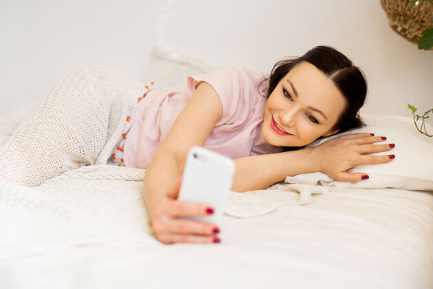 互联网卧室里漂亮的年轻女子拿着手机躺在床上手机早晨床