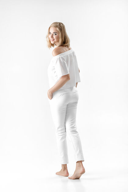 头发美丽微笑的金发女人在白色的背景上穿着白色的衬衫和裤子交叉金发向上