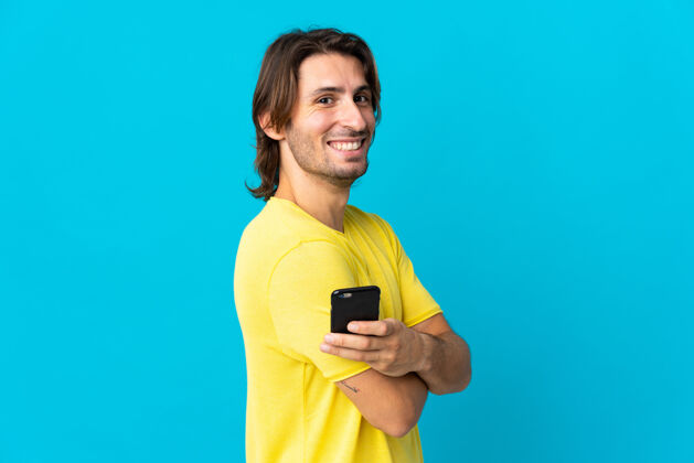 瘦身一个年轻英俊的男人被隔离在蓝色的墙上 手里拿着手机 双臂交叉手机肖像电话